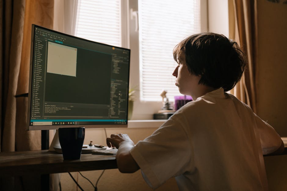 A person with short dark hair sits at a wooden desk indoors, working on a desktop computer with a large monitor displaying lines of code or programming interface. The individual is viewed from the sid